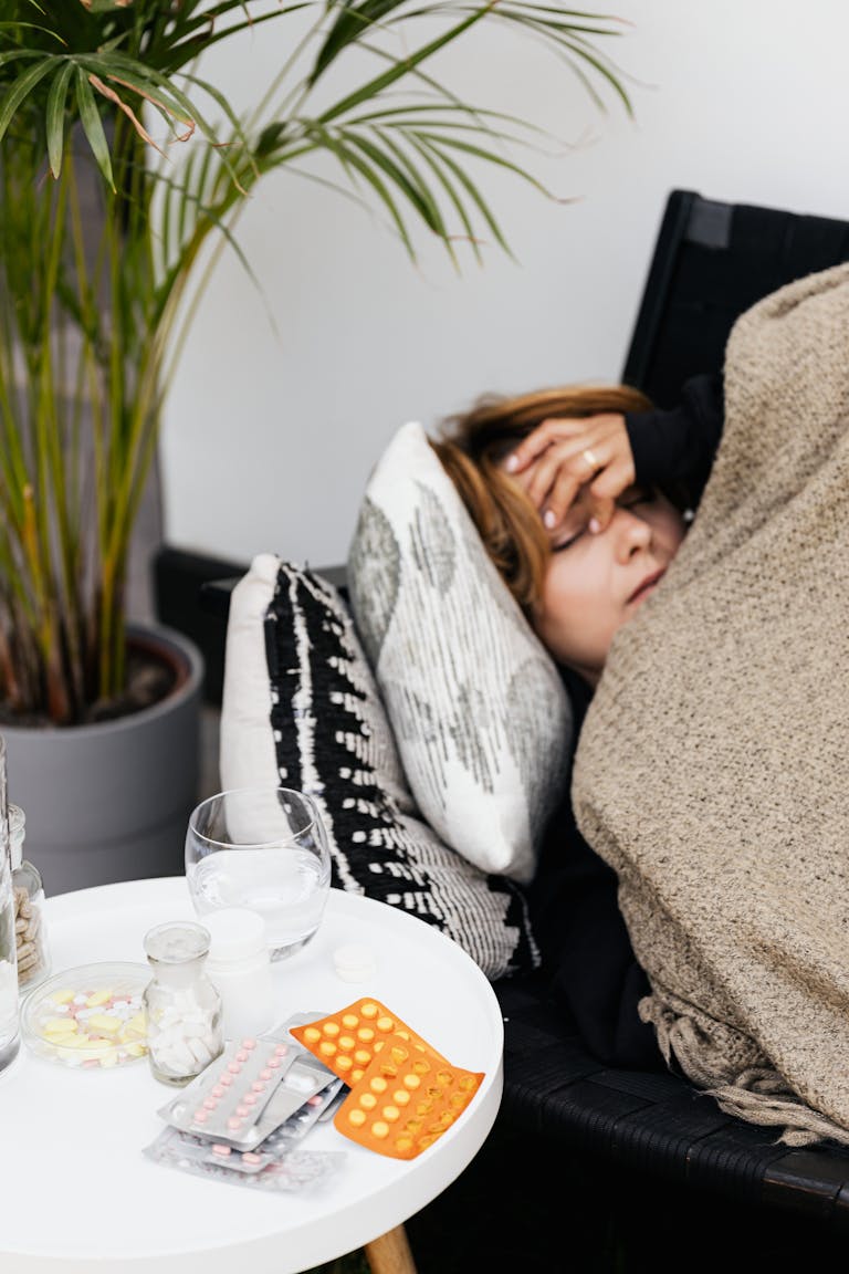 Woman lying on a couch covered with a blanket, feeling unwell, with pills on a table nearby.